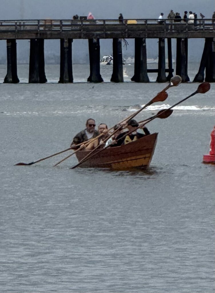 Photograph taken from a distance of four people seated in a wooden sewn-plank tomol Chumash canoe, paddling with long double-sided paddles across a harbor with a wharf in the background.