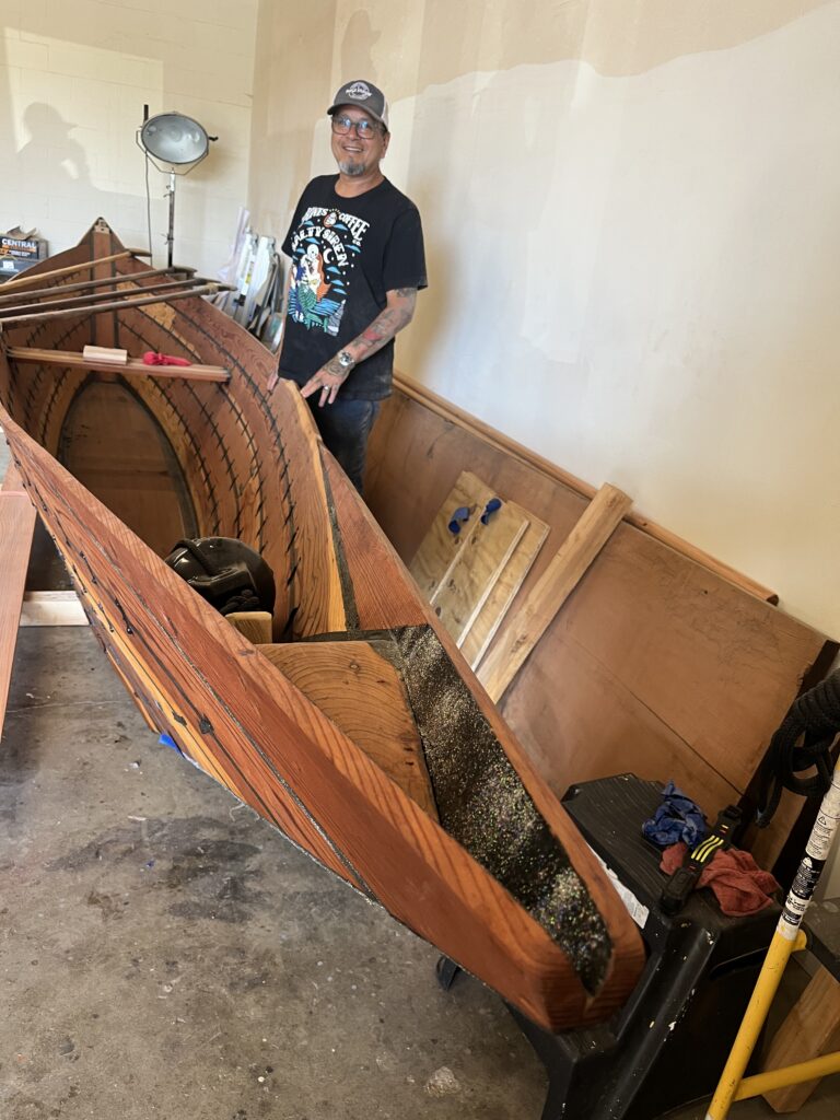 Photograph of the interior of a room featuring a man standing besides a freshly completed wooden sewn-plank Chumash tomol canoe sitting on wooden blocks.