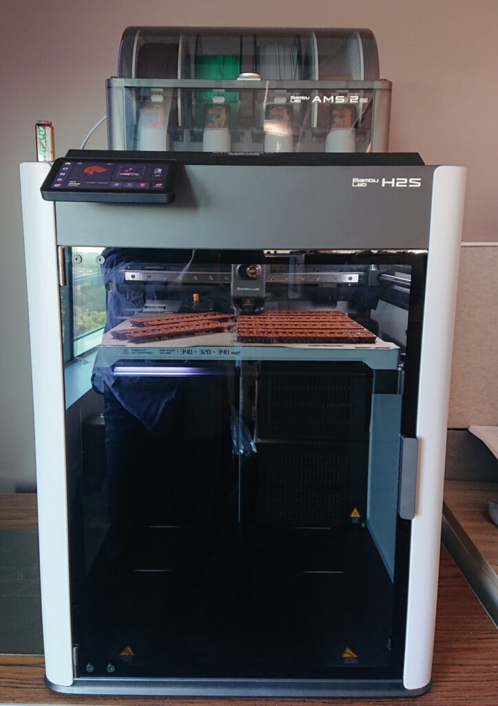 Photograph of a 3D printer with sheets of brown plastic logs being created.