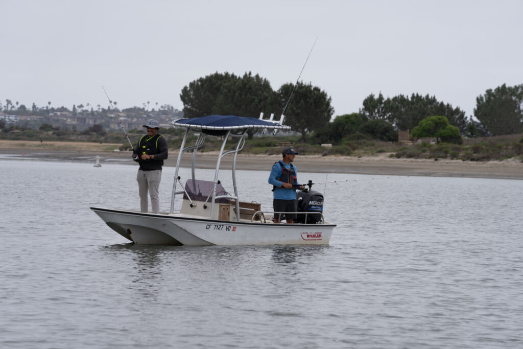 Two men aboard a small craft one fishing off the bow, the other off the stern in a tidal inlet.