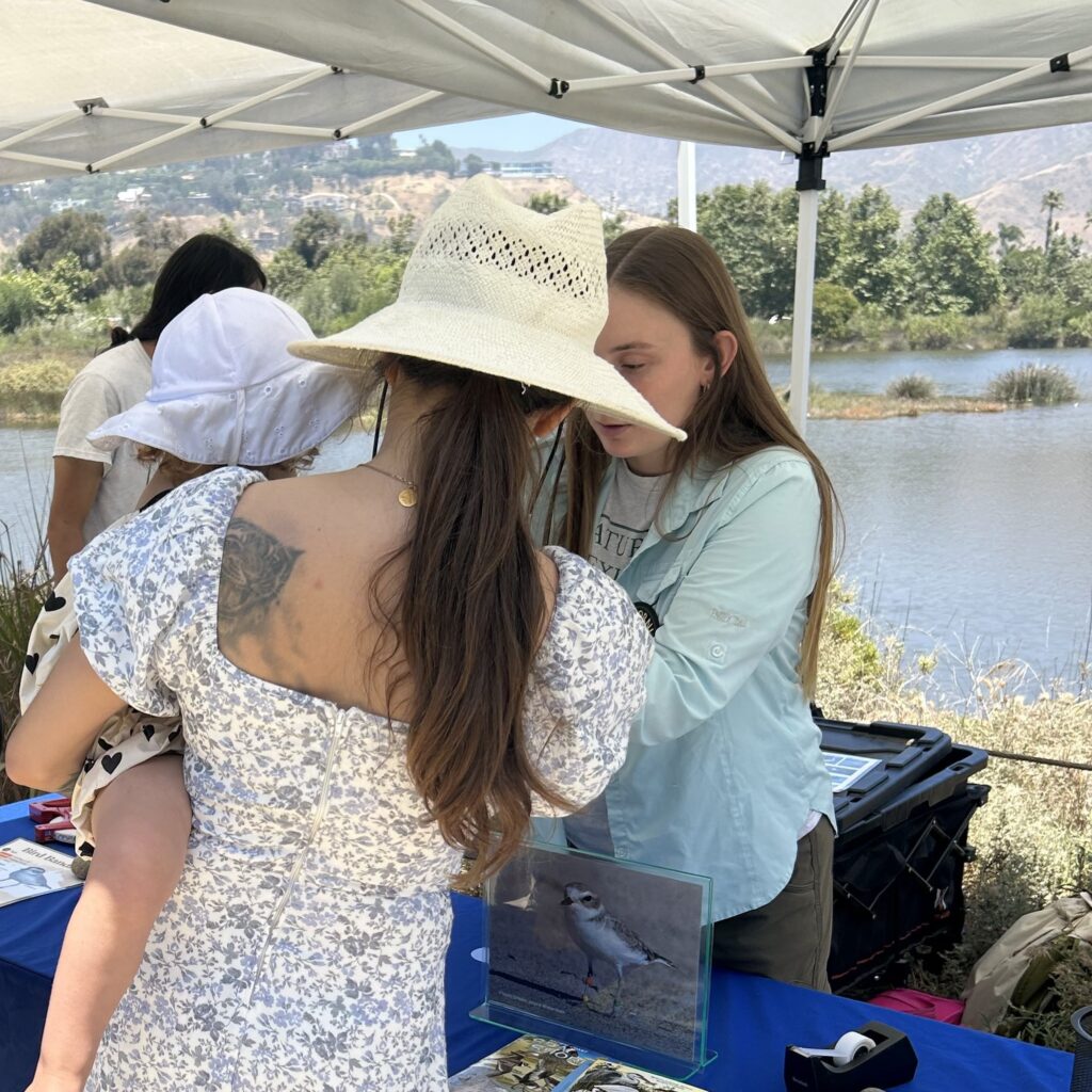 Photograph of a woman holding a child in her arms while speaking over an informational table to another woman, with Malibu Lagoon visible in the background.