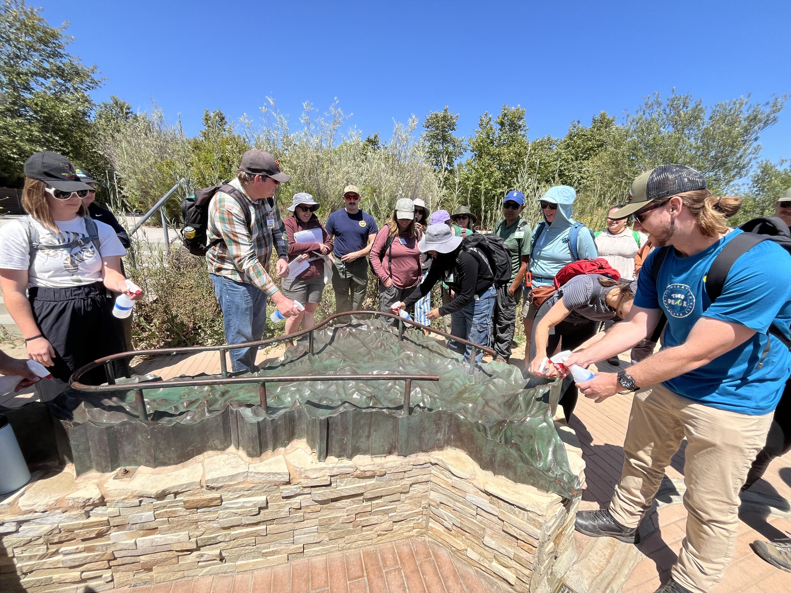 Mounted outdoor model of watershed mountains and valleys surrounded by training participants, some with spray bottles used to simulate rainfall above the scaled mountains.