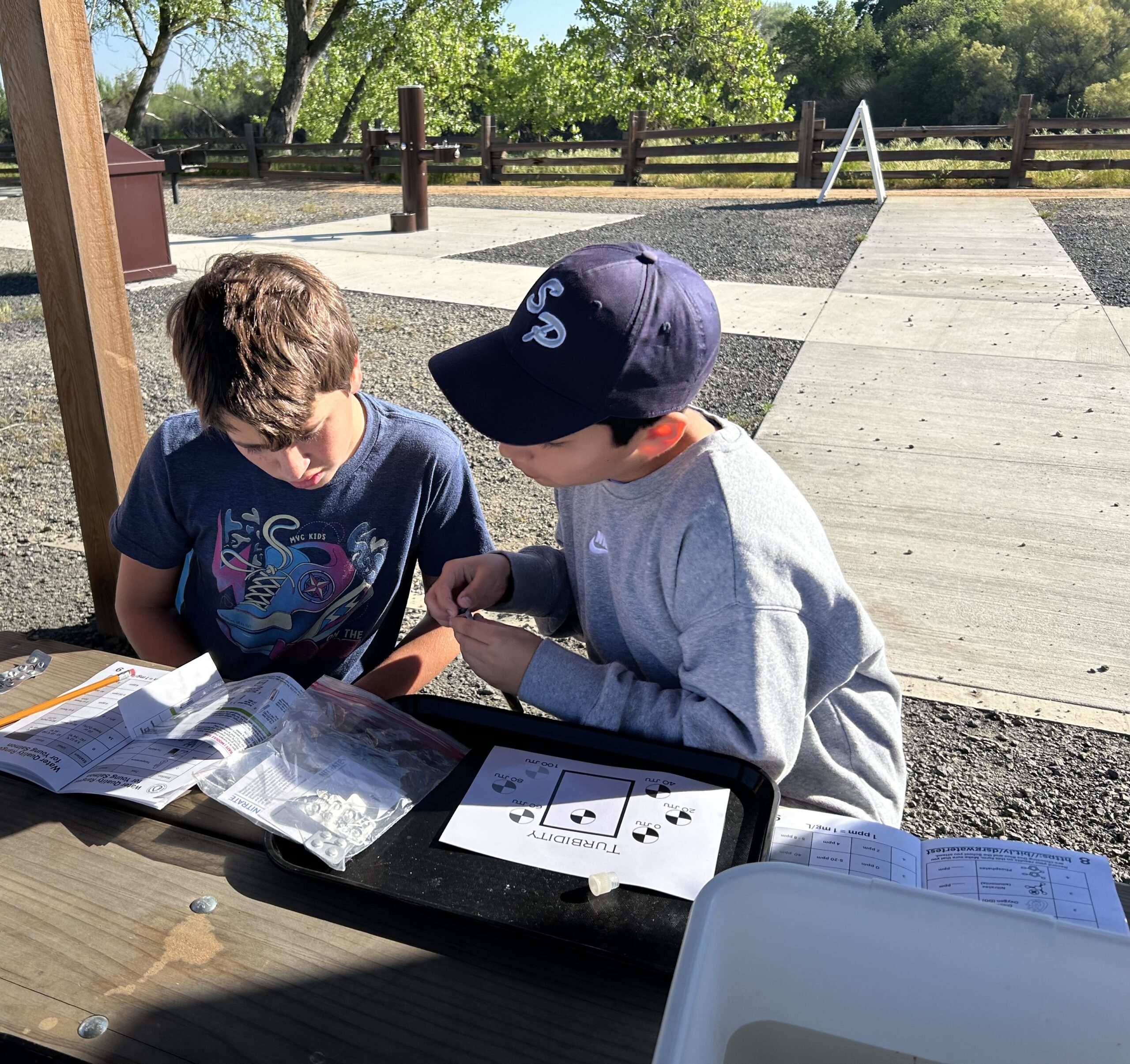Two young boys working together at an outdoor table with trees in background. 