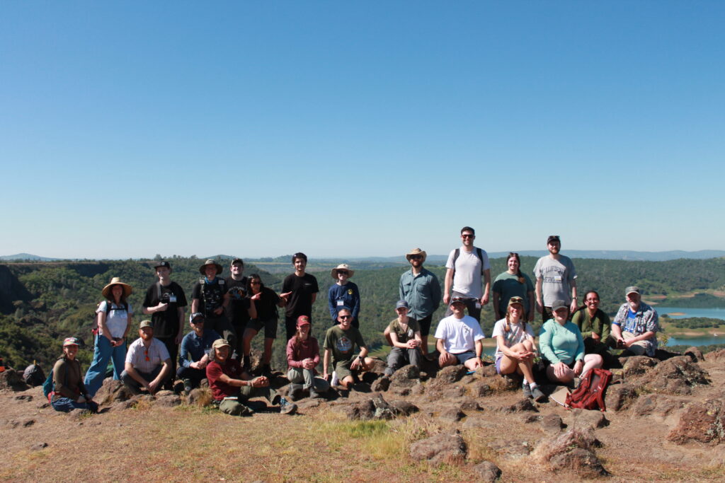 Photograph of a group of people wearing hiking clothing posing atop a ridgeline overlooking a background of rolling hills.