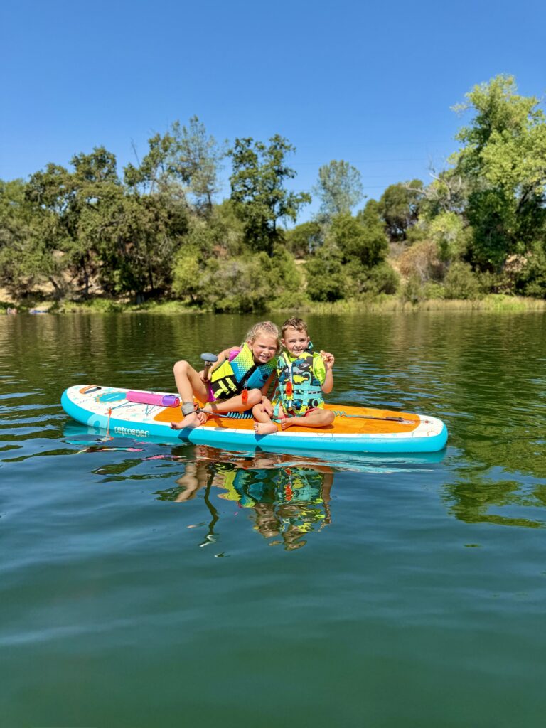 Photograph of two children wearing life vests enjoy a sunny day as they float while seated on a paddleboard on Lake Natoma.