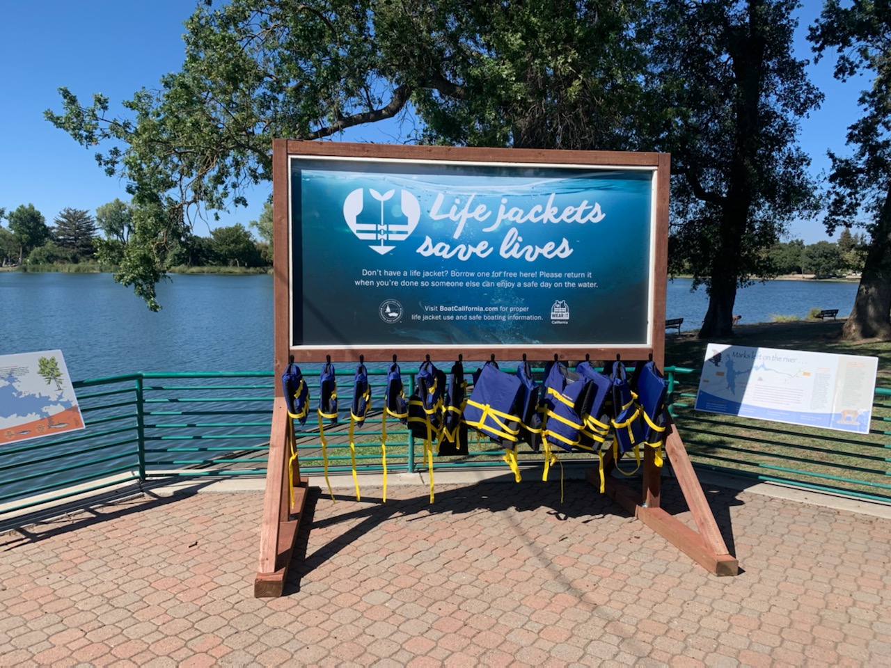 Rack supporting hanging life jackets of various sizes displays a sign that reads "Life Jackets Save Lives" standing above inland waterway.