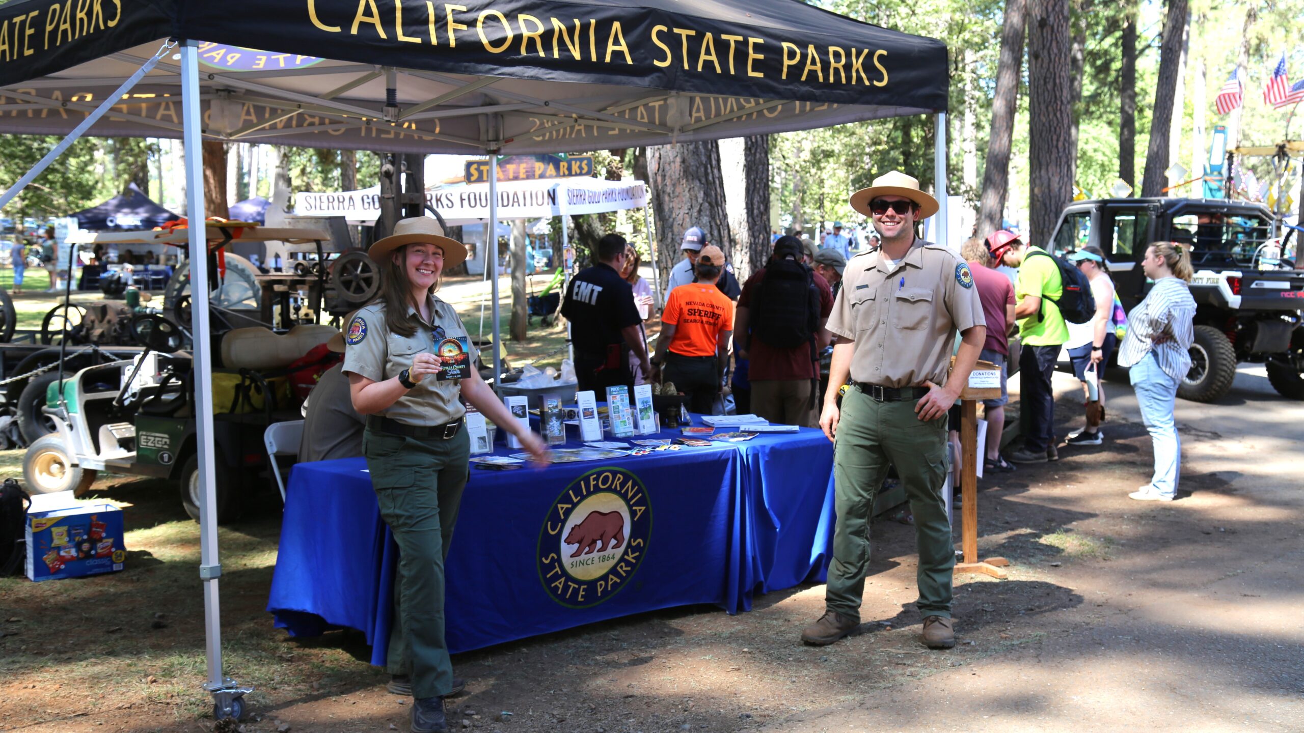 Photograph of two uniformed California State Park Interpreters standing in front of an outreach table at a forested fair grounds.