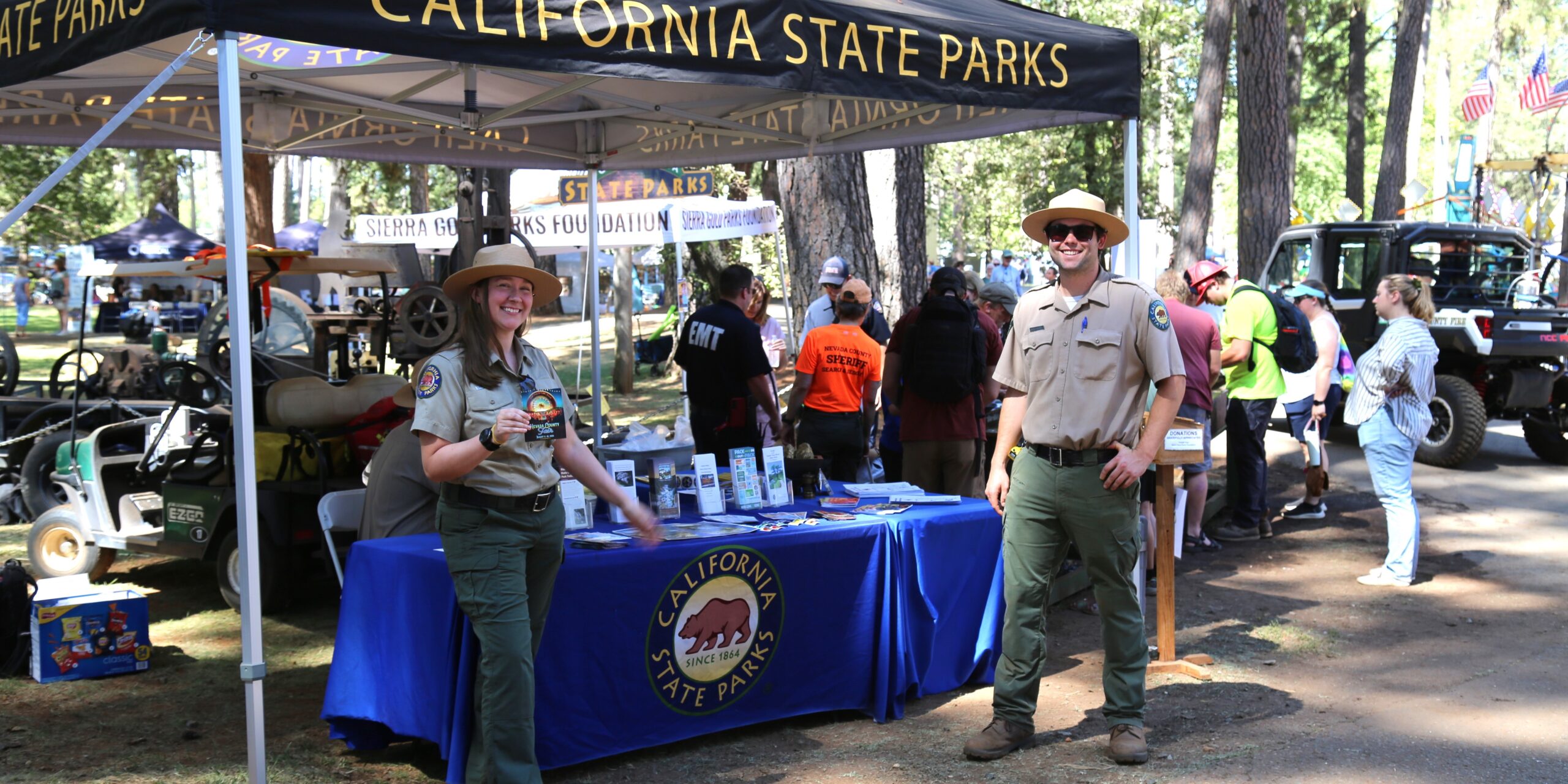 OHV Nevada Co Fair 2x1 Photograph of two uniformed California State Park Interpreters standing in front of an outreach table at a forested fair grounds.