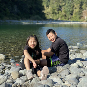 Two fourth grade students are smiling and sitting on a rocky bank. There is a calm, shallow river in the background.